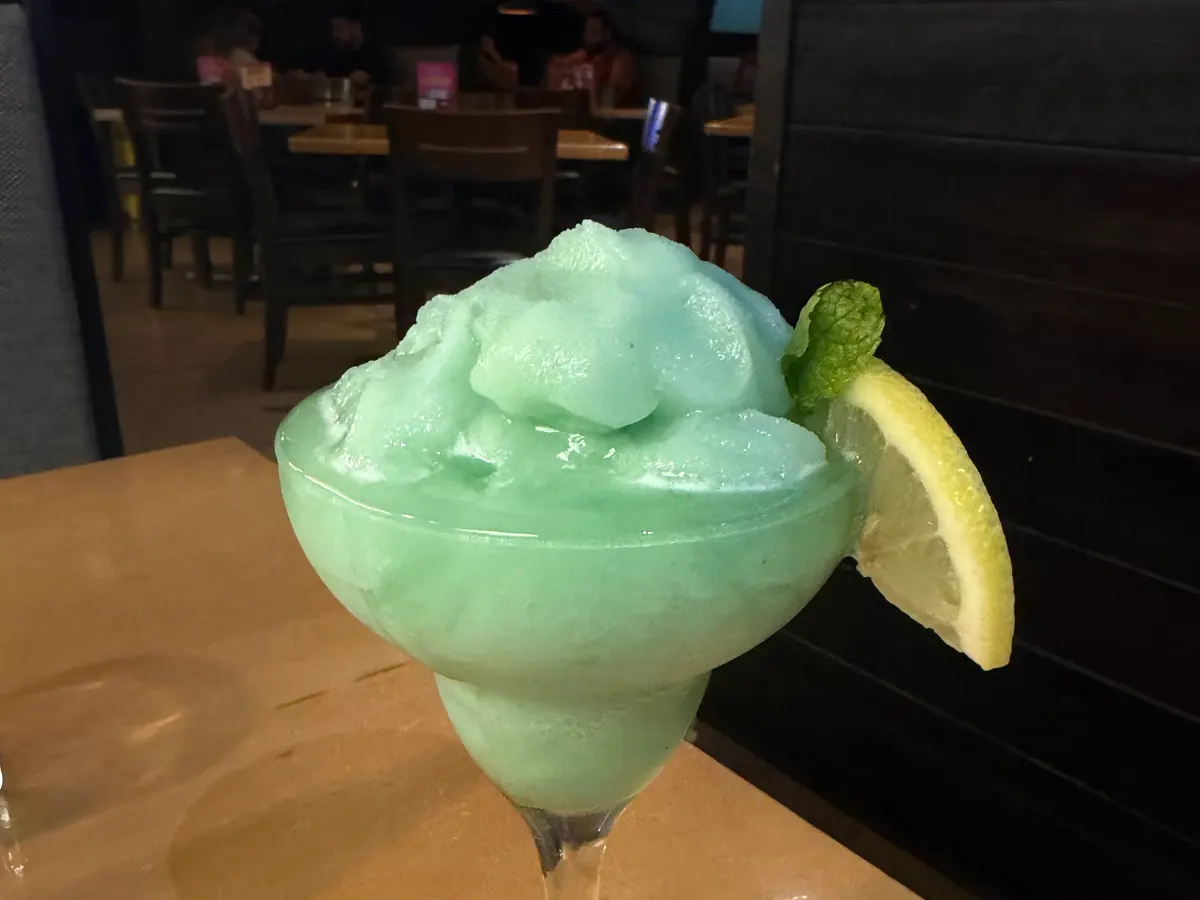 A light green frozen drink in a stemmed glass, with a lemon slice and a mint leaf on the rim